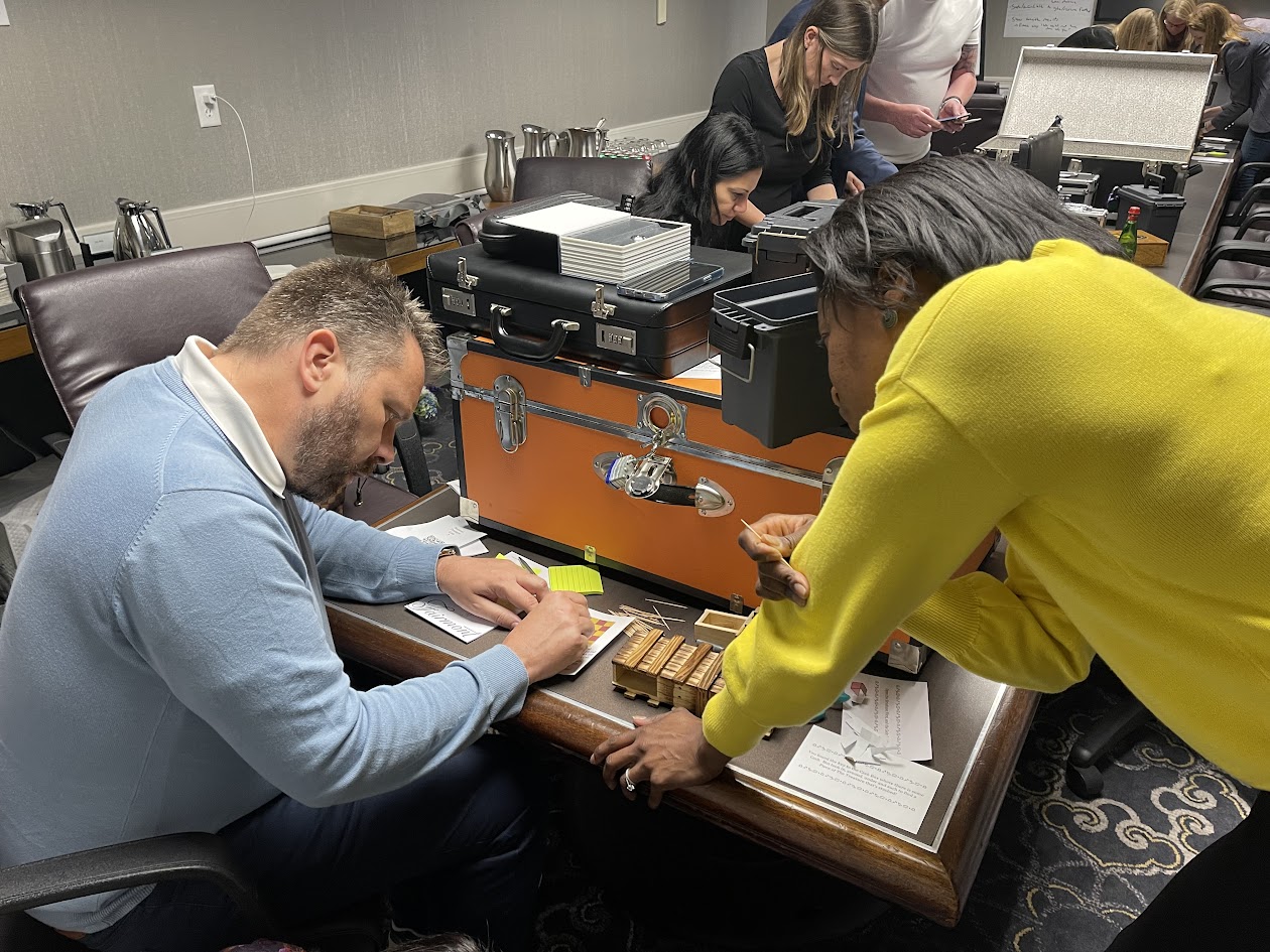 Two participants concentrate on solving a puzzle box using tools and a lock mechanism during a mobile escape room activity, with an orange lockbox on the table