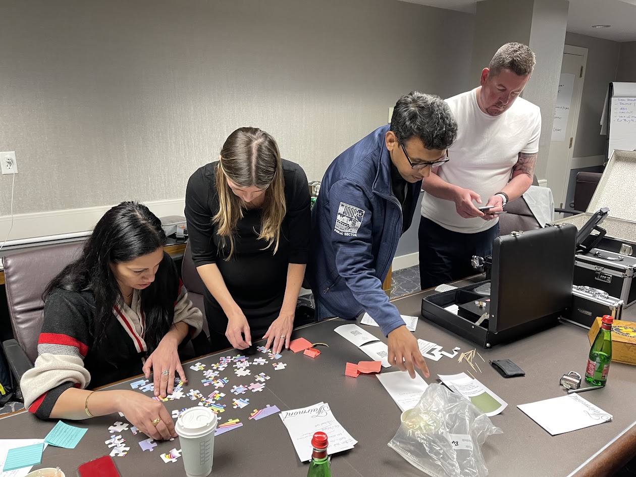 Four participants work together to assemble a colorful jigsaw puzzle on a table with a locked briefcase and clue cards during a mobile escape room event