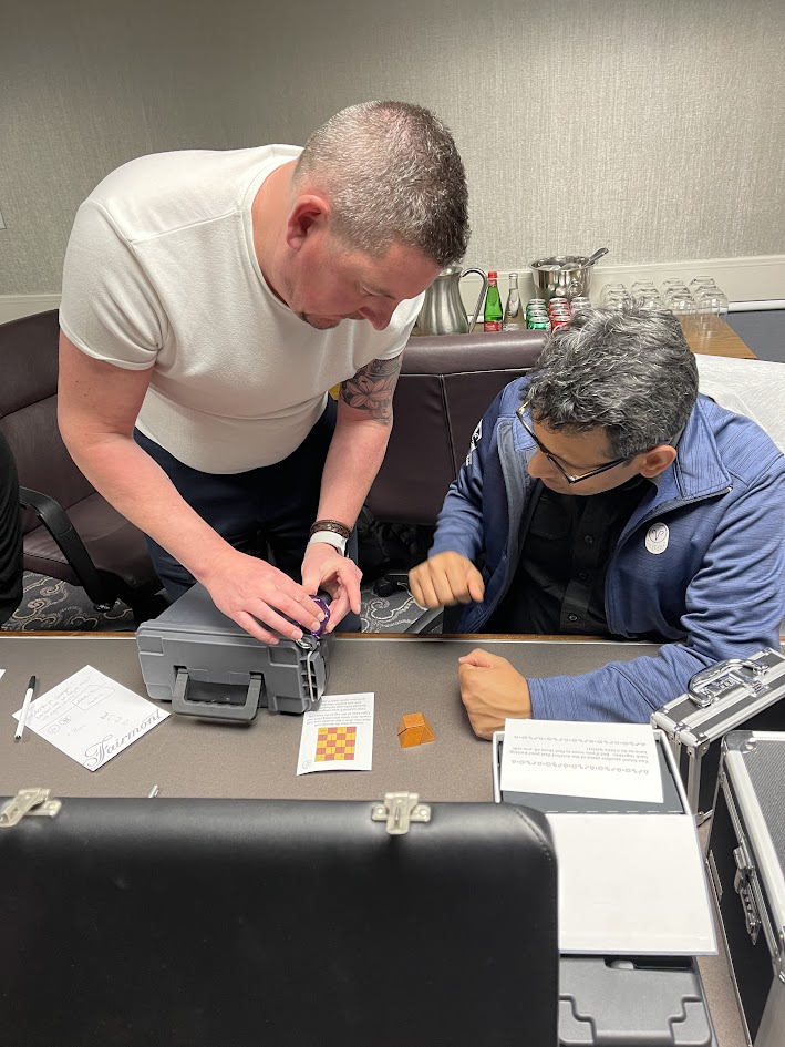 Two men collaborate closely on a small mechanical device and clues on a table during a mobile escape room challenge