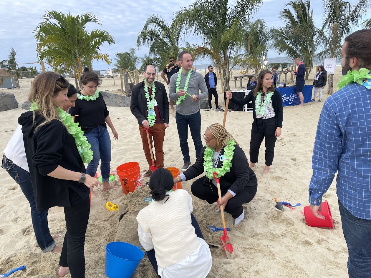 Team members wearing green leis work together to dig in the sand with shovels and buckets under palm trees on a beach