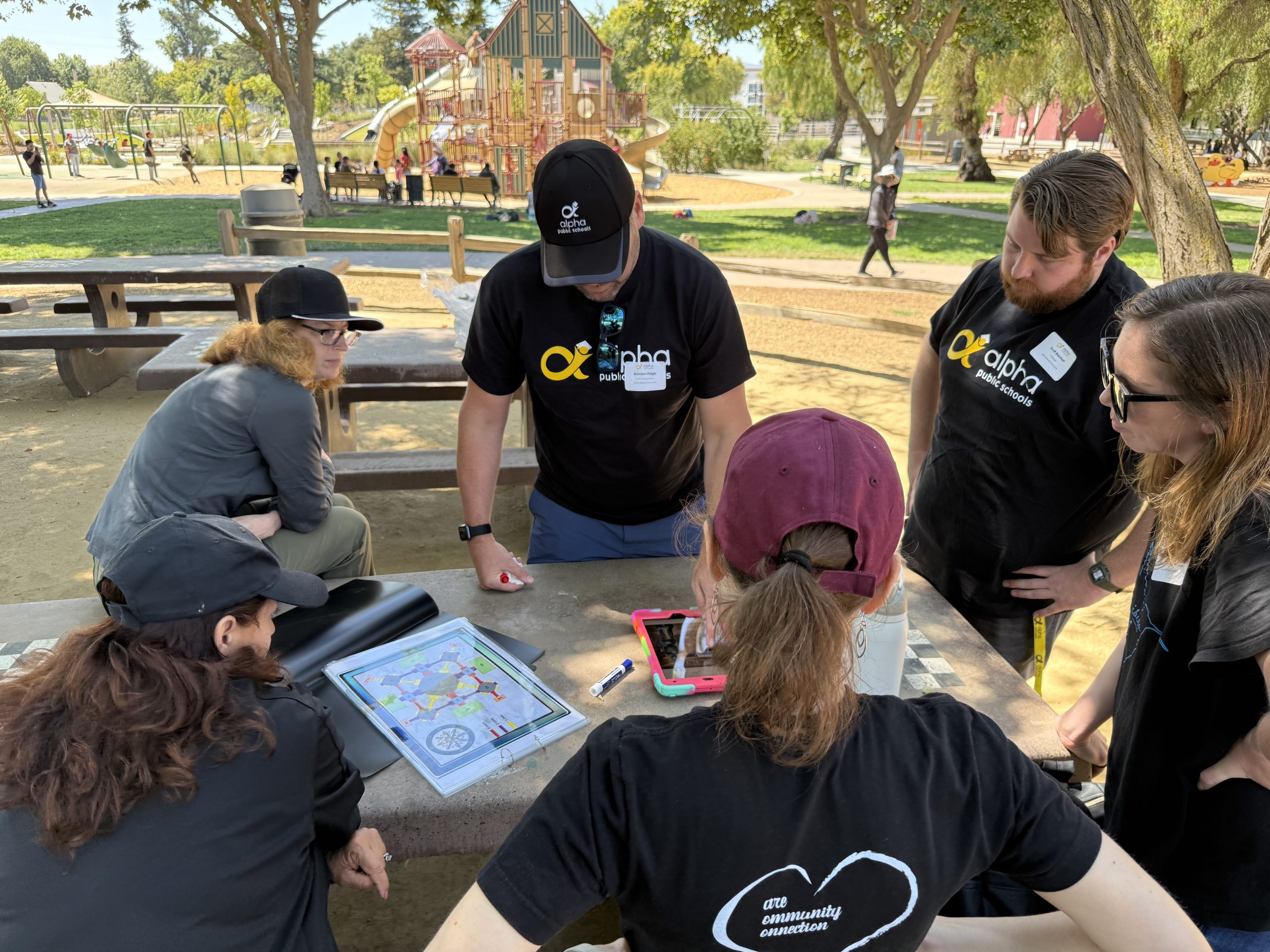 A group of people wearing matching apparel gathers around a park picnic table to collaborate on a planning project.