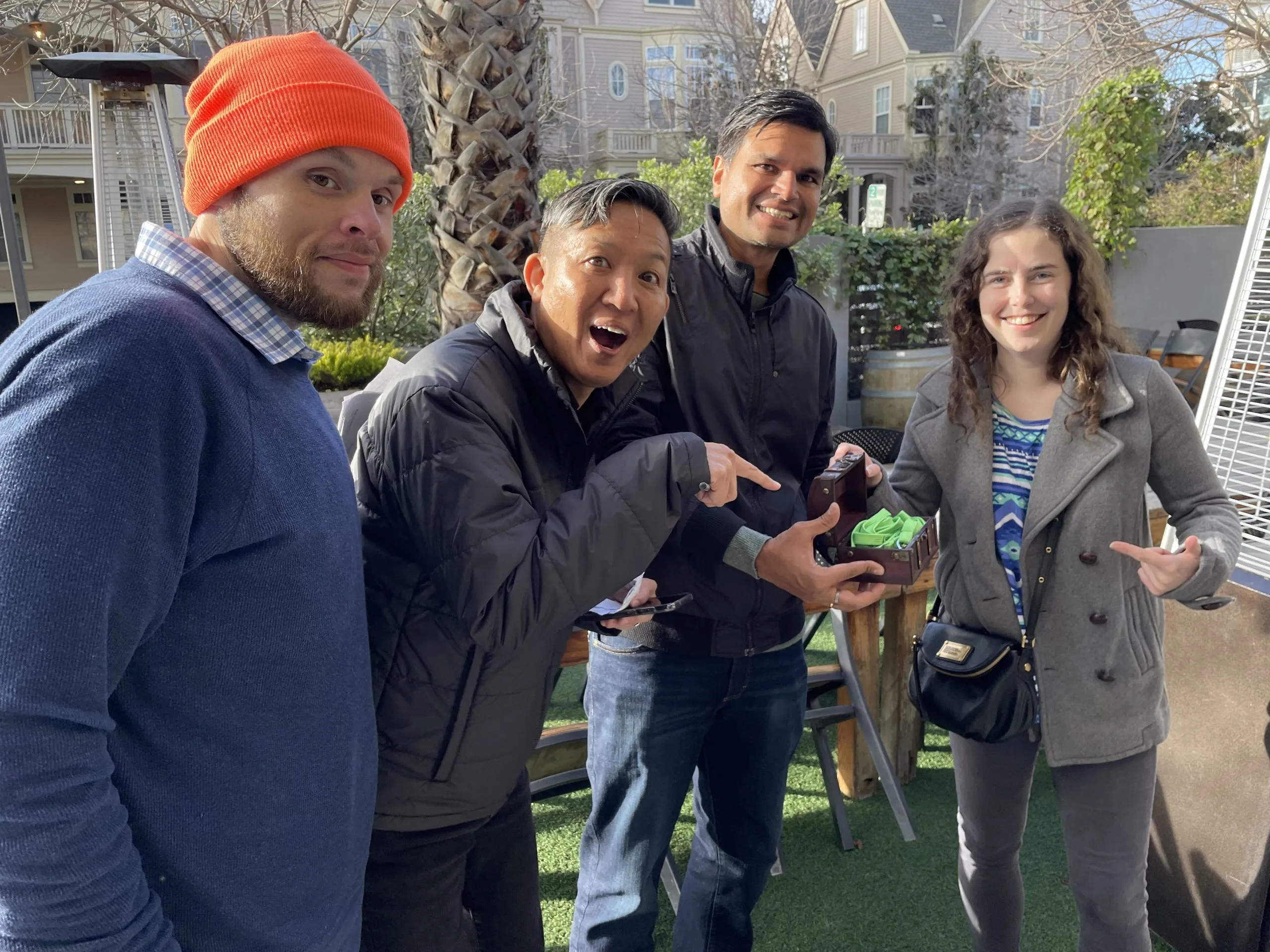 Four participants smiling outdoors in a courtyard—one wearing an orange beanie, another pointing excitedly, a man in a dark jacket, and a woman holding a green device