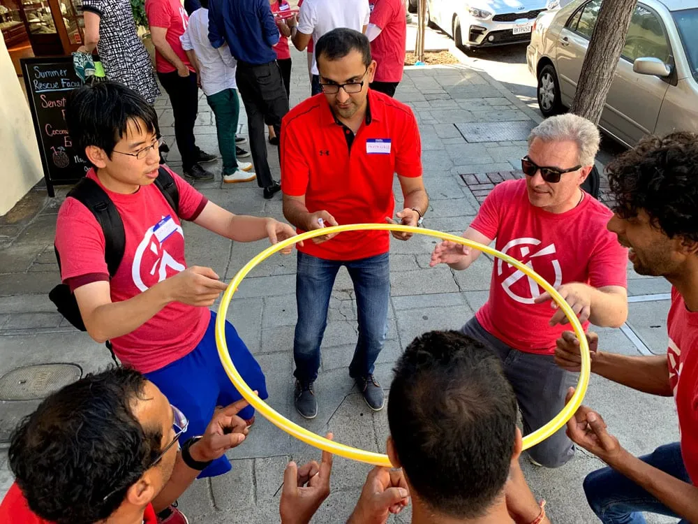 Team in matching red shirts attempting helium hula hoop challenge, passing yellow hoop hand-to-hand without letting go.
